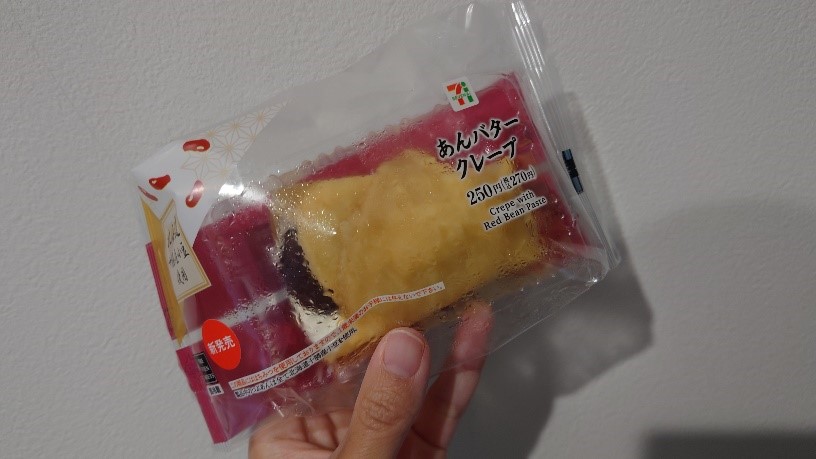 セブンイレブンの「7プレミアム あんバタークレープ」を手に持っている様子。パッケージには赤いデザインと商品の写真が見える。