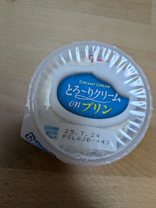 木目調のテーブルの上に置かれたグリコの「とろ~りクリーム on プリン」。蓋には25.7.24と印字された賞味期限が記載されている。