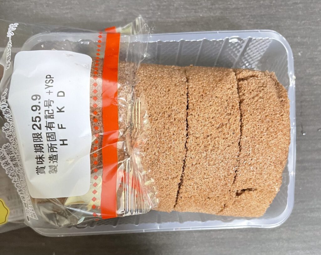 透明なプラスチックの容器に入った、切り分けられたチョコレートロールケーキの写真。ケーキは茶色い生地で、複数枚にスライスされ、側面に白いクリームが見える。容器の上には「賞味期限」と書かれた白いラベルが貼られている。