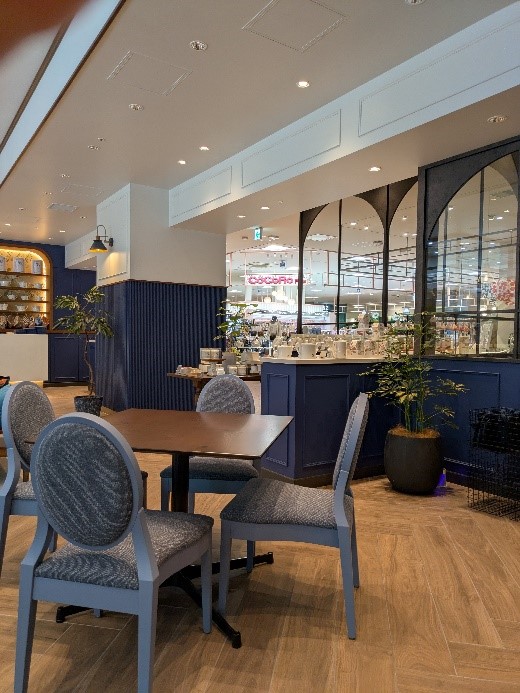 深いブルーを基調としたカフェの店内。木目の床と丸テーブル、クッション付きの椅子、観葉植物が配置され、落ち着いた雰囲気が漂う。