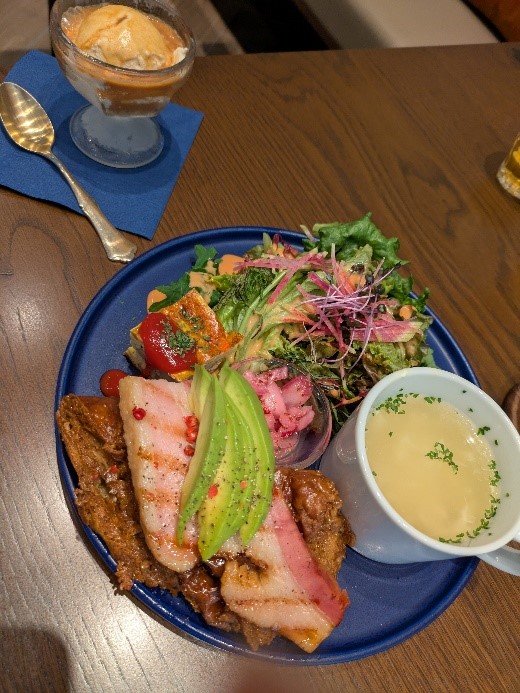 青いプレートに盛られたカフェランチ。厚切りベーコンとアボカド、サラダ、スープが並び、奥にはアイスクリームデザートが添えられている。