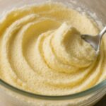 透明なガラスのボウルに入った、泡立てられた淡い黄色のクリーム。スプーンがクリームをすくい上げ、なめらかな質感と泡立ちを示している。